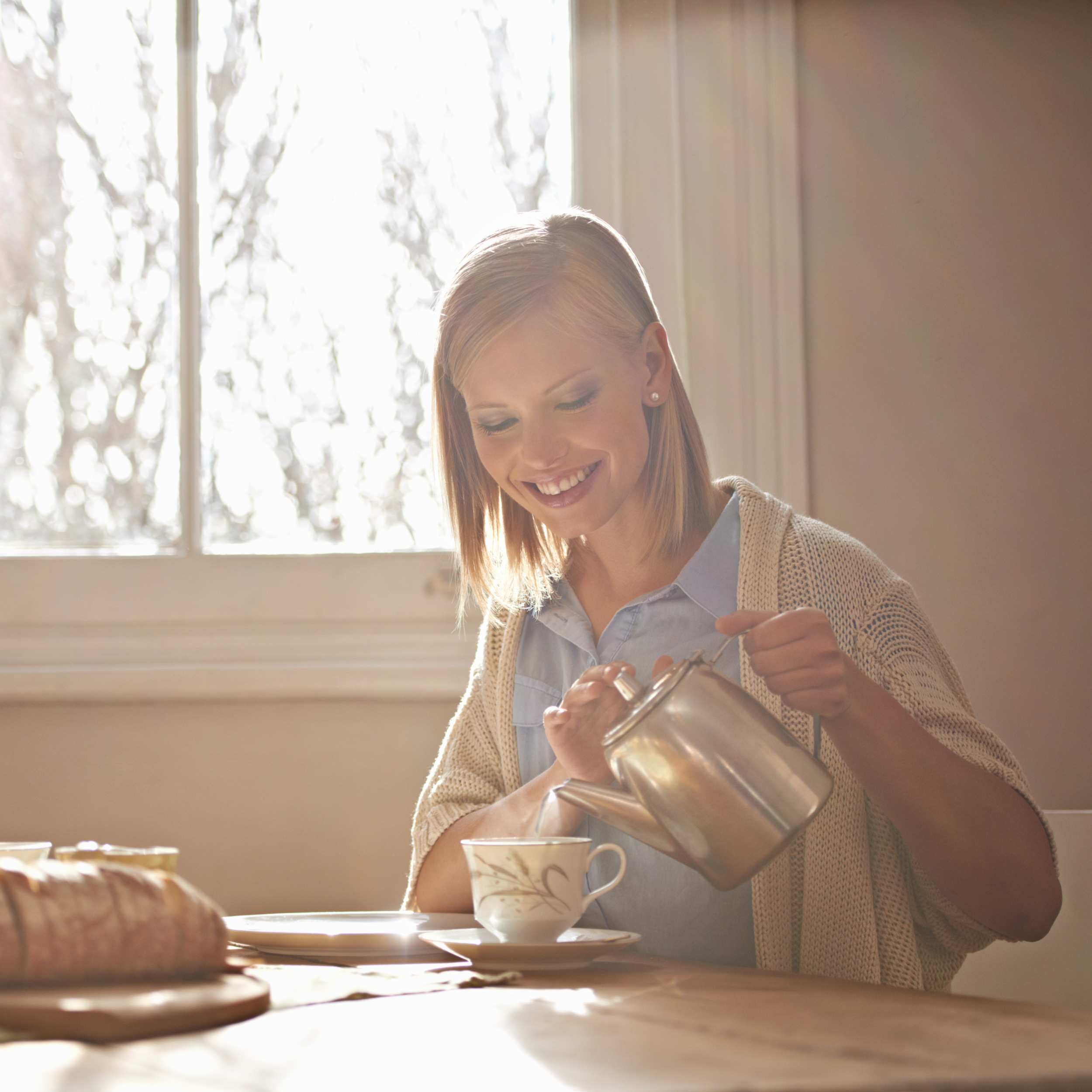Pouring, tea and happy woman in home for morning, calm routine and drink on table. Girl, relax and coffee break with food, snacks or teapot in hands for breakfast preparation on holiday or vacation.
